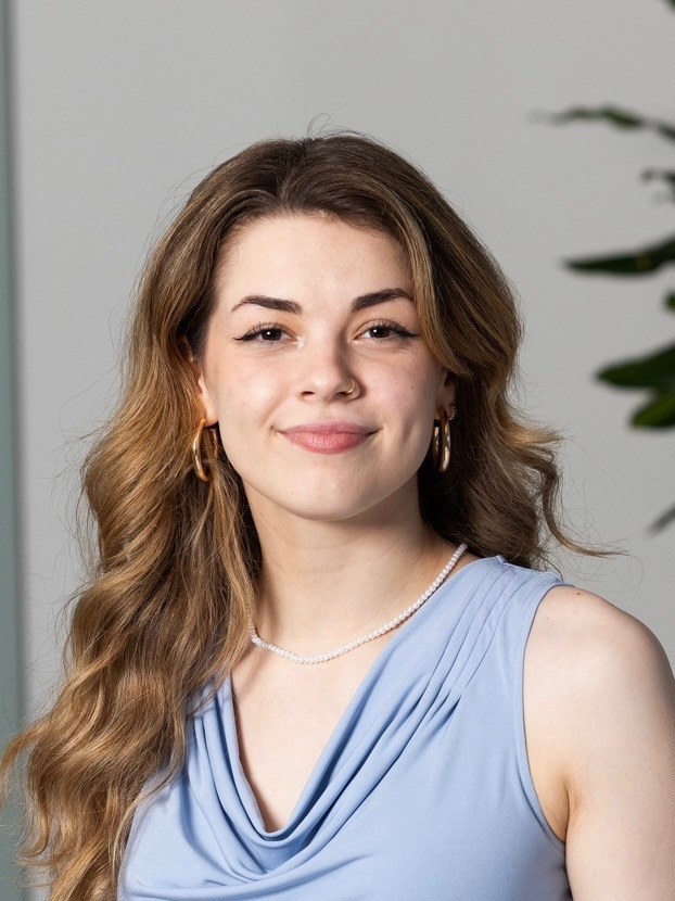 Emily Weinberg, a young woman with long wavy light brown hair, wearing a pale blue top and a thin pearl necklace, smiling gently at the camera.